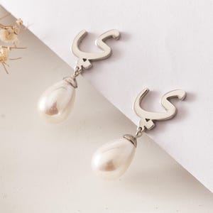May include: A pair of silver-toned earrings featuring a stylized Arabic letter design. Each earring has a teardrop-shaped, lustrous white pearl pendant suspended below the letter. The earrings are displayed on a white surface.