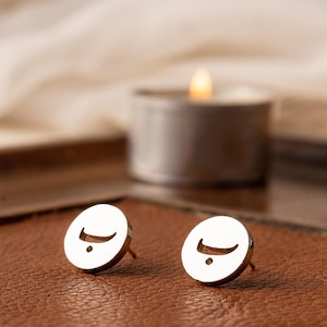 May include: A pair of silver-colored stud earrings. Each earring features a circular design with a curved line and a small dot. The earrings are displayed on a brown surface, with a lit candle in the background.