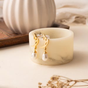 May include: Gold-colored earrings with a crescent moon design and dangling teardrop-shaped pearls. The earrings are displayed on a marble-patterned candle holder. A lit candle and dried flowers are in the background.