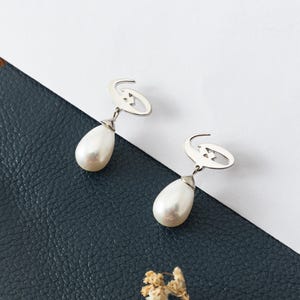 May include: A pair of silver-toned earrings with a teardrop-shaped pearl pendant. The earrings have a unique design with a curved top and a small star-like detail. The background is a mix of white and dark blue.