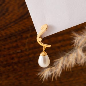 May include: A gold-colored earring with a stylized design, featuring a teardrop-shaped pearl pendant. The earring is attached to a white surface, set against a blurred brown background with dried plants.
