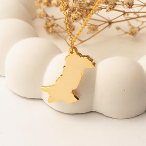 Puede incluir: Un collar dorado con un colgante con forma de mapa de Pakistán. El colgante cuelga de una delicada cadena dorada. El collar se exhibe sobre una pieza decorativa blanca en forma de nube, con flores secas en el fondo.