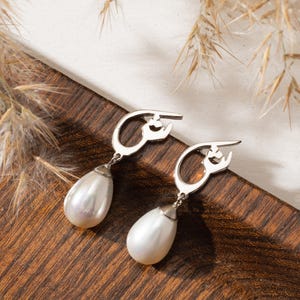 May include: A pair of silver earrings with a teardrop-shaped pearl pendant. The earrings are displayed on a wooden surface with dried plants in the background. The pearls have a lustrous, white appearance.