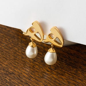 May include: A pair of gold-colored earrings with a unique design. Each earring features a stylized gold shape with a teardrop-shaped, faux pearl pendant. The earrings are displayed on a wooden surface.
