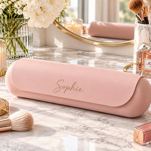 Puede incluir: Un estuche de almacenamiento cilíndrico de color rosa claro con un acabado mate suave. La parte frontal del estuche presenta el nombre Sophie impreso en una delicada escritura cursiva dorada. El estuche está colocado horizontalmente sobre una superficie de mármol blanco.