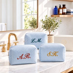 May include: Three light blue waffle-textured toiletry bags with embroidered initials. One bag features dark green L.M. initials, another has red A.R. initials, and the third displays gold S.K. initials. Each bag has a zipper closure and a rectangular shape.