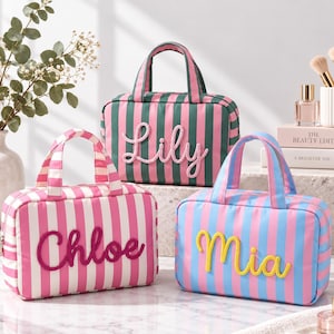 May include: Three rectangular cosmetic bags with top handles and vertical stripes. One bag is pink and white with the name Chloe in dark pink, one is green and pink with Lily in pink, and one is blue and pink with Mia in yellow script lettering.