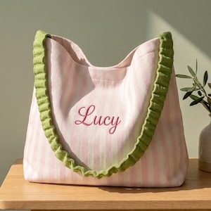 Op de afbeelding: Een tote bag met een verticaal gestreept patroon in lichtroze en wit, met de naam Lucy geborduurd in donkerroze cursief schrift. De tas heeft twee geplooide olijfgroene stoffen hengsels die aan de bovenrand zijn bevestigd.