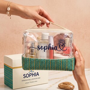 May include: A clear rectangular toiletry bag with a teal and gold woven base, featuring the name Sophia in dark blue script with a sun and wave icon. The bag contains various cosmetic bottles and is paired with a matching white gift box.