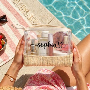 Peut inclure: Une trousse de toilette transparente avec une base en paille tressée et une fermeture éclair. Le devant de la trousse porte le prénom Sophia en lettres cursives noires suivi d'un cœur. Divers produits de beauté sont visibles à l'intérieur.