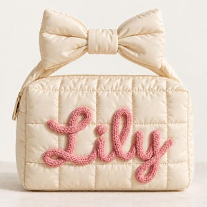 May include: A cream-coloured quilted rectangular pouch featuring a large bow handle on top. The front is decorated with the name Lily in pink knitted yarn lettering. The bag includes a gold-toned zip pull on the side for secure closure.