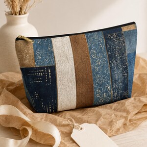 May include: A zippered pouch featuring a vertical patchwork design of textured fabric panels in shades of navy blue, cream, and brown. The blue sections are decorated with subtle, light-colored botanical and geometric line art patterns.