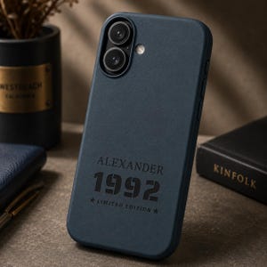 May include: A dark blue phone case with a matte texture featuring the embossed text ALEXANDER, the number 1992, and the words LIMITED EDITION flanked by two small stars. The case has a dual-lens camera cutout and a circular flash opening at the top.
