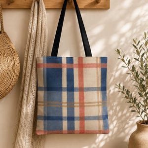 May include: A rectangular tote bag featuring a plaid pattern with intersecting blue, coral pink, and beige stripes. The bag has two solid black fabric shoulder straps attached to the top edge, providing a contrast to the light-colored plaid body.