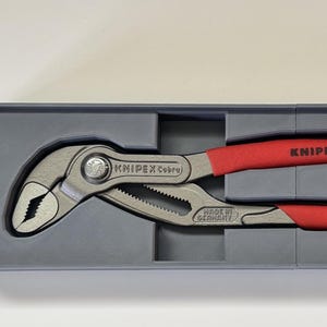 May include: A pair of Knipex Cobra pliers with red handles, presented in a gray box. The pliers are silver with a toothed gripping surface. The red handles have the text "KNIPEX" and "87 01 250".