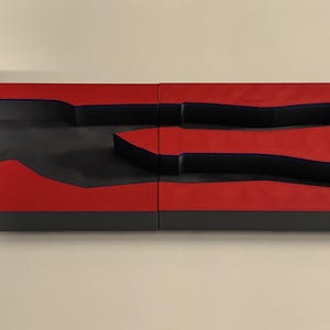 Op de afbeelding: Een tweedelige, rechthoekige mal met een rode bovenkant en zwarte zijkanten. De mal heeft een menselijke figuur en twee andere vormen die in het bovenoppervlak zijn uitgesneden. De mal is ontworpen voor het maken van objecten.