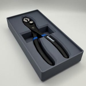 Peut inclure: Une pince noire et bleue, marquée "KOBALT", présentée dans un étui rectangulaire gris. La pince a des accents bleus sur les poignées et est conçue pour saisir et couper. L'étui est une solution de rangement.