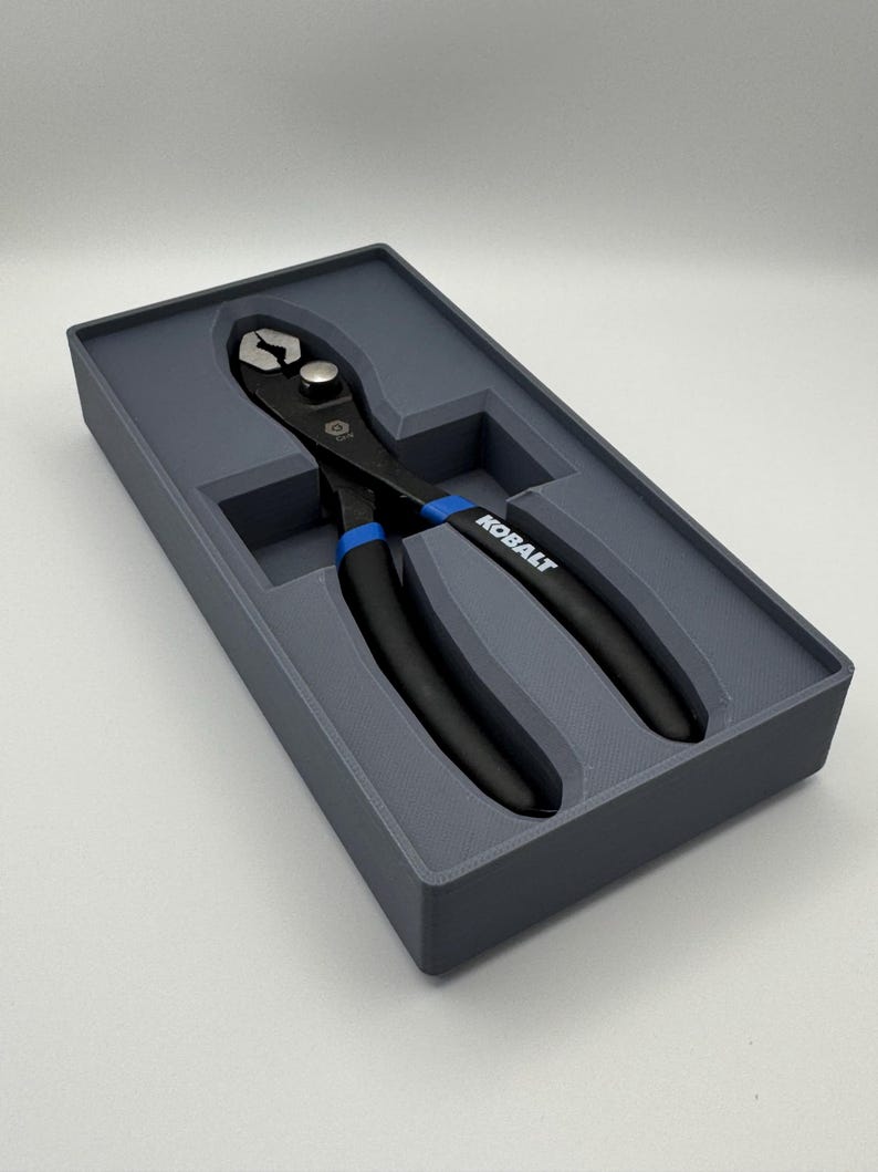 Op de afbeelding: Een zwarte en blauwe tang, met de merknaam "KOBALT", in een grijze, rechthoekige koffer. De tang heeft blauwe accenten op de handgrepen en is ontworpen om te grijpen en te snijden. De koffer is een opbergoplossing.