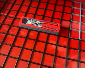 May include: A pair of pliers with red handles and silver jaws, resting in a blue and red box. The pliers are in a red toolbox with a black grid pattern.