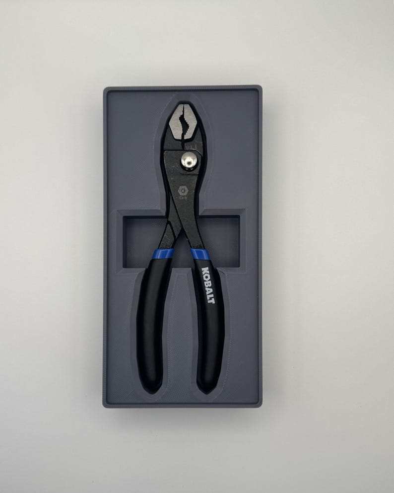 Op de afbeelding: Zwarte waterpomptang met blauwe handgrepen, gepresenteerd in een grijze tray. De tang heeft een zilveren draaipunt en het woord "KOBALT" is op de handgreep gedrukt. Deze tangen zijn ontworpen voor het grijpen en manipuleren van objecten.