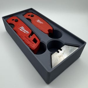Peut inclure: Un organisateur en plastique gris contient deux cutters rouges Milwaukee FASTBACK et une lame de cutter argentée. Les couteaux sont dans un plateau en plastique gris avec des découpes circulaires. Le logo Milwaukee est visible.
