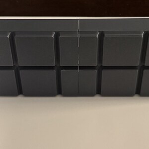 Op de afbeelding: Een donkergrijs, rechthoekig object met een reeks verhoogde, vierkante blokken. De blokken zijn in een rasterpatroon over het oppervlak gerangschikt. Het object lijkt van een massief materiaal te zijn en heeft een matte afwerking. Ongeveer 20 cm lang.