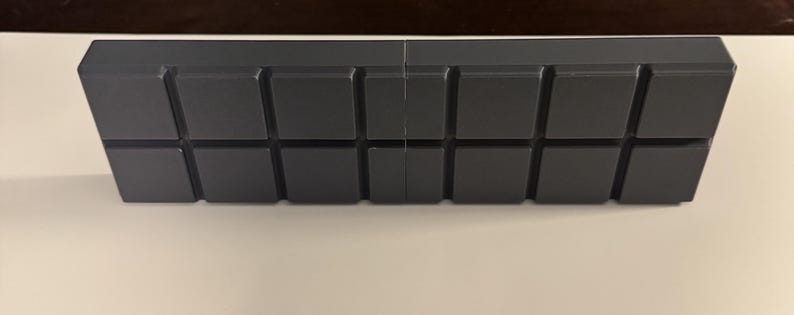 Op de afbeelding: Een donkergrijs, rechthoekig object met een reeks verhoogde, vierkante blokken. De blokken zijn in een rasterpatroon over het oppervlak gerangschikt. Het object lijkt van een massief materiaal te zijn en heeft een matte afwerking. Ongeveer 20 cm lang.
