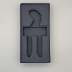 K&ouml;nnte beinhalten: Eine dunkelgraue rechteckige Schale mit geformten F&auml;chern. Die Schale hat einen gebogenen oberen Abschnitt, einen rechteckigen mittleren Abschnitt und zwei vertikale Schlitze unten. Das Design deutet darauf hin, dass es zum Organisieren oder Halten von Gegenst&auml;nden dient.