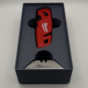 Peut inclure: Un cutter rouge avec la marque "Milwaukee" et le texte "FASTBACK" imprimé, présenté dans une boîte gris foncé. Une lame argentée est également visible.