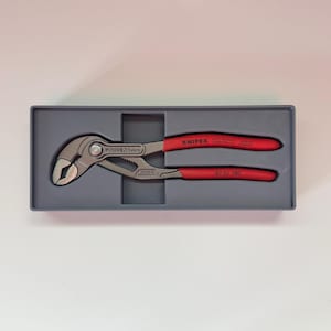 May include: A set of Knipex Cobra pliers in a gray case. The pliers are silver with red handles. The text "KNIPEX" is printed on the handle. The pliers are designed for gripping and turning objects.