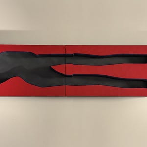 Op de afbeelding: Een tweedelige, rechthoekige, rode houten doos met een zwarte, mensvormige uitsparing. De uitsparing is een gestileerde weergave van een liggende persoon. De doos staat op een wit oppervlak.