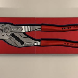 Op de afbeelding: Een Knipex tang met rode handgrepen, gepresenteerd in een rode doos. De tang is zilver en zwart, met de merknaam en het modelnummer zichtbaar. De doos is open en toont het gereedschap.
