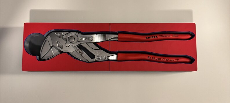 Op de afbeelding: Een Knipex tang met rode handgrepen, gepresenteerd in een rode doos. De tang is zilver en zwart, met de merknaam en het modelnummer zichtbaar. De doos is open en toont het gereedschap.