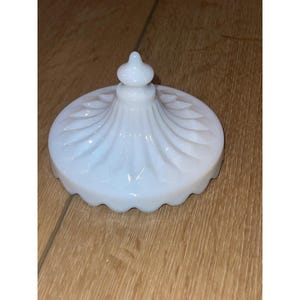 May include: A white, opalescent glass lid with a decorative, scalloped edge. The lid features a central finial and a swirling, textured pattern radiating from the center. The lid is likely for a jar or container.