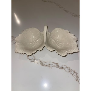 May include: A white, leaf-shaped serving dish with two compartments and a handle. The dish has a textured surface with detailed veining, and is made of ceramic or porcelain. The design is symmetrical and elegant, suitable for serving snacks or small portions.
