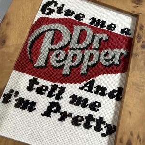 May include: A cross-stitch embroidery in a light brown wooden frame featuring the text "Give me a Dr Pepper And tell me I'm Pretty" in black and silver thread against a white background with a large red oval shape behind the brand name.