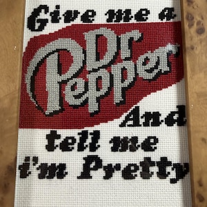 May include: A cross-stitch embroidery in a light brown wooden frame featuring the Dr Pepper logo in silver and black against a red background, with the black text "Give me a Dr Pepper And tell me I'm Pretty" stitched on a white fabric grid.