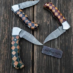 May include: Three folding pocket knives with Damascus steel blades and handles featuring a diamond-patterned inlay of wood and colored resin. A small rectangular metal tag with the text Soul Forge Crafts is positioned near the knives.