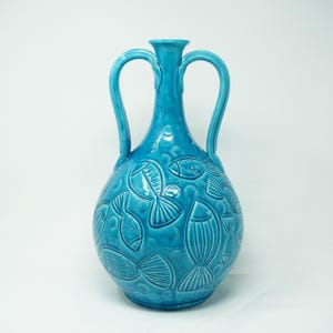 May include: A turquoise ceramic vase with two handles and a detailed fish pattern. The vase has a bulbous body and a narrow neck. The fish are embossed on the surface, along with bubbles.