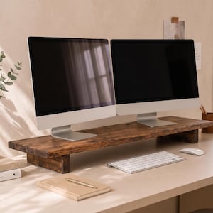 May include: Two silver-framed computer monitors sit on a dark brown wooden monitor stand. A white keyboard and mouse are in front of the stand. A vase with greenery and a small potted plant are on the left.