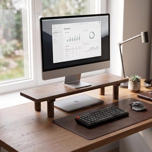 May include: A wooden monitor stand with four cylindrical legs sits on a wooden desk. A silver computer monitor displays a productivity dashboard. Below the stand is a closed silver laptop, a black mechanical keyboard, a gray mouse, and a brown leather desk mat.