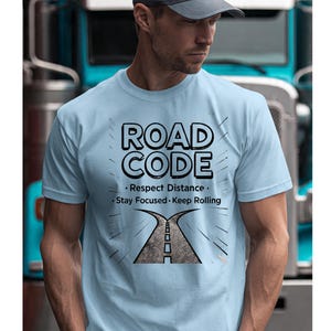 Può includere: T-shirt azzurra con la scritta "ROAD CODE" in grassetto nero. Sotto, le frasi "Rispetta la distanza", "Rimani concentrato" e "Continua a rotolare". Un grafico di una strada che si divide in due è sotto.