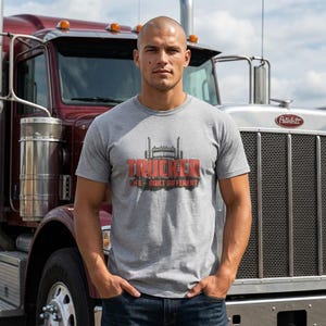 Könnte beinhalten: Ein graues, meliertes Kurzarm-T-Shirt mit einer Grafik der Vorderseite eines Sattelschleppers, dem roten Schriftzug TRUCKER und dem kleineren schwarzen Text LIFE - BUILT DIFFERENT auf der Brustpartie.