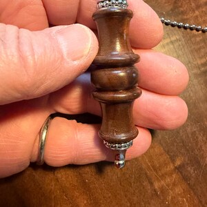 May include: A brown wooden pendulum with silver-colored metal accents. The pendulum has a cylindrical shape with a rounded top and bottom, and is suspended from a silver chain. The top and bottom of the pendulum are decorated with silver metal.