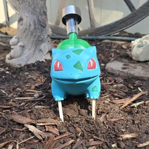 May include: A light blue and green Bulbasaur garden stake featuring a solar-powered light fixture attached to the back. The creature has red eyes and dark green spots, supported by two thin white stakes inserted into the dark brown soil.