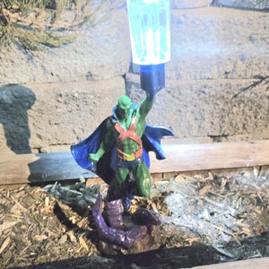 Puede incluir: Una figura verde de Martian Manhunter con una capa azul y una lámpara solar. La figura sostiene la lámpara por encima de su cabeza. La base de la figura es de color púrpura oscuro.
