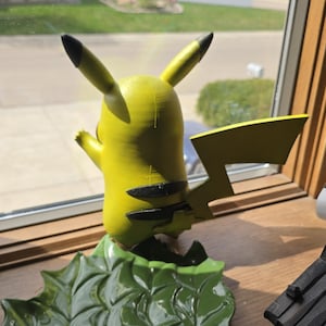 Peut inclure: Figurine jaune de Pikachu avec des détails noirs sur les oreilles et le dos, posée sur une base verte en forme de feuille. La figurine est placée près d'une fenêtre, avec une vue sur des maisons et de la verdure en arrière-plan.