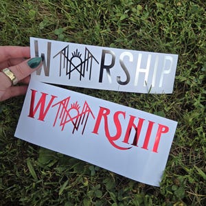 May include: Two rectangular vinyl decals on white backing paper. Each decal features the word WORSHIP in a stylized font, with the letter O replaced by a geometric symbol featuring a sunburst and vertical lines. One decal is silver, the other is red.