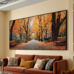 May include: A large framed canvas print depicting an autumn scene. A brick pathway is lined with trees displaying vibrant orange and yellow foliage. Benches and street lamps are visible along the path. The artwork is displayed above a brown sofa with decorative pillows.