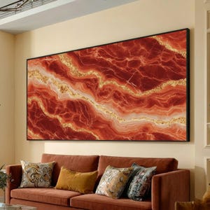 Puede incluir: Gran obra de arte mural rectangular con marco negro. La obra presenta un diseño abstracto vibrante en tonos rojos, dorados y blancos, que se asemeja a una formación geológica. La obra cuelga sobre un sofá marrón.